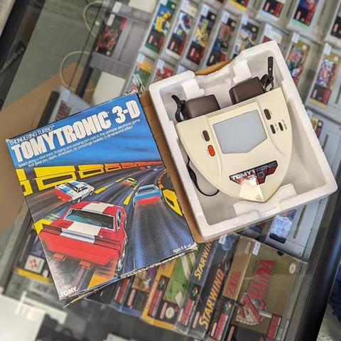 Close-up of a vintage Tomytronic 3‑D “Thundering Turbo” handheld game in its original box and styrofoam packaging. The cream-colored binocular-style device sits beside its illustrated box showing cars racing along a curved highway.