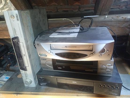 One black and two silver VHS decks stacked. A silver DVD player standing on its narrow side left of the others. All ar a bit dusty.