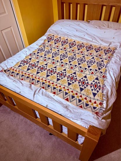 A handmade crochet blanket lies neatly across a wooden bed with a white duvet. The blanket features a repeating geometric pattern of diamonds and triangles in navy blue, mustard yellow, red, and cream yarn. Its edges are fringed, and the bed frame is light oak with vertical slats. The room has warm yellow walls, a beige carpet, and a white door to the left.