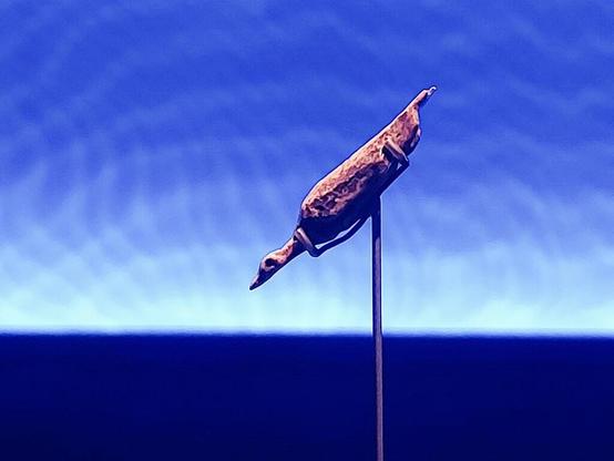 A photo of a small bird figurine mounted on a thin metal rod, set against a blue gradient background. The figurine appears angled downward, with a pointed head and elongated body, creating a simple abstract silhouette.