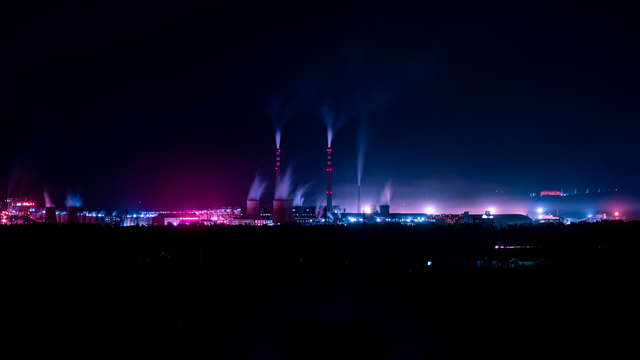 A night photo of a power plant or other industrial facility on the horizon, with smoke and steam rising out of huge chimneys. The blues and purples are very exagerated on the photo