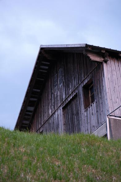 a barn clad in washed out, grey-ish wooden planks, sat atop a grassy elevation, photographed from below against an overcast but bright sky