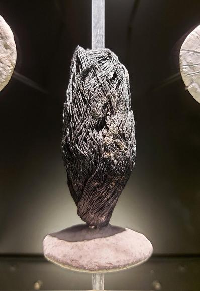 A charred skein of linen thread displayed upright in a museum case. The blackened, twisted fibers are tightly coiled and partially fused from burning, mounted between two smooth, pale stone supports. The exhibit is lit to highlight the texture and preservation of the ancient textile remains.