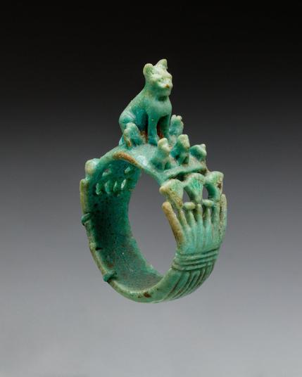 Turquoise-green ring with a sculpted cat perched on top and several small kitten figures grouped beside it, the band decorated with vertical ridges and floral-like raised ornamentation.