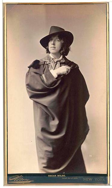 This photograph of Oscar Wilde in a dramatic pose, taken by Napoleon Sarony around 1882, shows him standing and facing slightly to the left. Wilde has medium-length wavy hair and wears a wide-brimmed fedora-style hat. He has a large dark cape over his shoulders and a light-colored cravat or neck scarf tied in a big bow. His right hand is lifted near his chest, holding the cape in place.