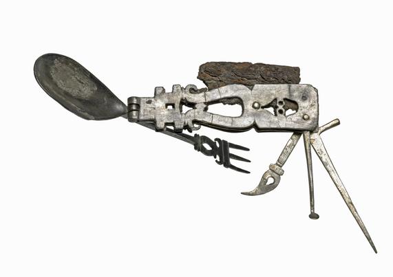 The folding multi-tool, it is built around a silver handle and a set of folding, pivoted implements. Its most visible feature is the broad oval spoon at the left; additional utensils fold from the opposite end and beneath the handle. A dark, heavily corroded piece along the top is the original iron knife blade.
