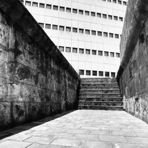 Black and white photograph of a stone-walled outdoor corridor leading to a short set of stairs. In the background is the façade of a mid-century international style office building featuring several rows of small square glass windows.