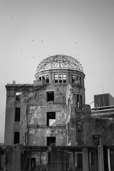 The Genbaku Dome, A-bomb Dome, Hiroshima Peace Memorial: a building standing after the 6th August 1945 atomic bombing of Hiroshima because it was almost directly below the blast. Birds are flying out of it. The roof is a dome of iron bar, complete and together but all gaps.