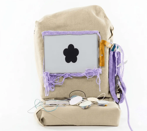 Se ve un extraño artefacto como hecho o forrado de saco, compuesto de una parte colocada en vertical,  y otra en horizontal. Pantalla blanca y negra en la parte vertical, enmarcada por borde tejido e hilos colganderos de color lavanda, y laterales con otros tejidos del mismo estilo y color

en la parte horizontal hay cuatro botones forrados de algún tejido.

Se ve blandito y se extrapola que es una especie de ordenador
