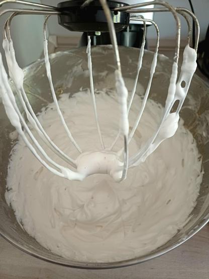 Aquafaba batida em chantilly em uma vasilha de metal.  Acima, a  pra da batedeira, com chantilly.