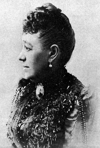 Jane Stafford appears in profile, shown from the chest up and facing left. Her dark hair is styled in an updo with curls on top and the rest pulled back. She wears a dark, textured dress with a cameo brooch at the center. The dress features beadwork, sequins, and embroidery.