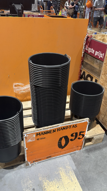 Een stapel bouwemmers op een palet. Op het schapbord staat “mannen handtassen”