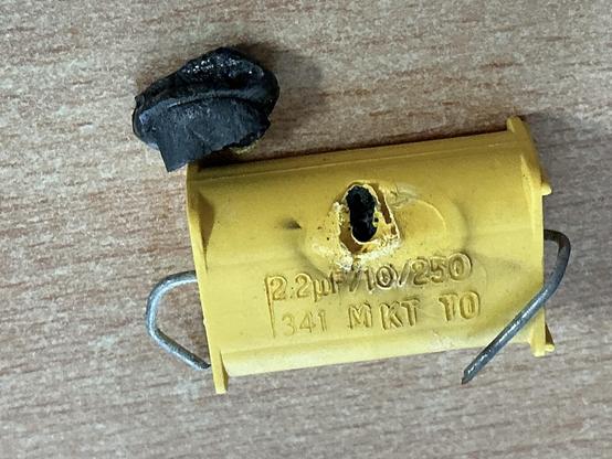 Foto toont een condensator waarvan het binnenwerk verbrand is en vloeibaar naar buiten was gelopen door een gat in de behuizing.