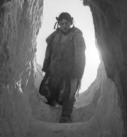 Photo of young Inuit woman staring ahead into the camera as she enters her igloo, walking down the steps carved from snow and ice. The walls of the structure are also made of packed snow. She's framed by the entrance, the Arctic sun shines through the opening in the door.

(Richard Harrington / Library and Archives Canada)