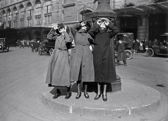 Three Parisian women watching the solar eclipse of 8 April 1921 through glasses on the Cour du Havre, next to the gare Saint-Lazare.

Three women in long coats stand on a raised platform beside a lamppost outside Gare Saint-Lazare, each holding improvised viewing devices to their eyes to observe a solar eclipse. Early automobiles and a busy street crowd fill the background.