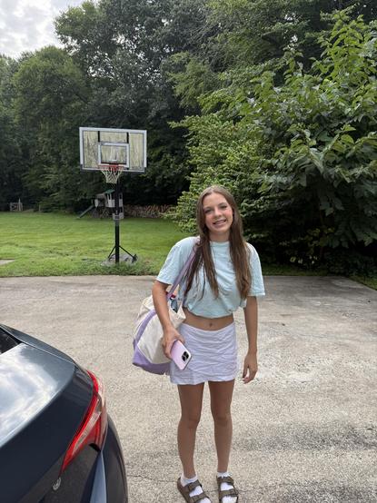 A smiling teenage girl with long brown hair stands on a driveway beside a car, wearing a light blue cropped T-shirt, white skirt, socks, and sandals. She carries a purple-and-white tote bag and holds a phone. A basketball hoop, green lawn, and dense trees fill the background.