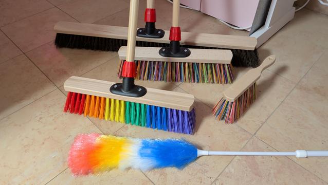 A bunch of brooms on my floor. The center one is the famous pride broom, surrounded by a colorful set of a hand broom and a hall broom, a duster and a big outdoor broom.