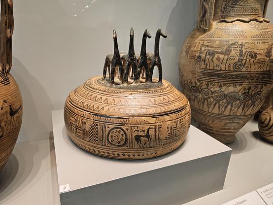 A horse pyxis displayed in a museum case: A low, round lidded terracotta container covered with dark geometric bands, zigzags, rosettes and animal scenes. Four stylised black horses stand in a row on the lid, their long necks rising above the vessel; larger decorated pottery jars are visible behind it.