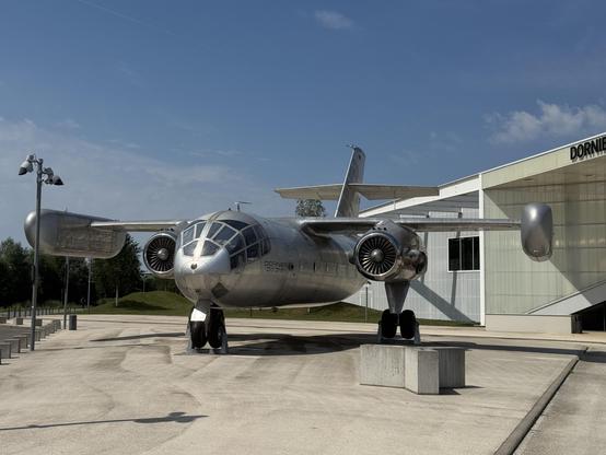 A bright sunny day with clear skies; concrete plaza in the foreground, museum building just visible behind it, and in the foreground, one of the only two Dornier Do-31 prototypes built—the world's only vertical take-off jet transport aircraft, powered by a Rolls-Royce pegasus engine (as on the Harrier) under each wing, with giant pods on the wing-tips that each contain four vertically-oriented smaller jet engines for take-off. The Do-31 is otherwise fairly mundane aside from the glazed cockpit and nose (a bit like a B-29) and being generally very silvery and futuristic … a future we didn't get, as the project was cancelled after test flights in 1971.