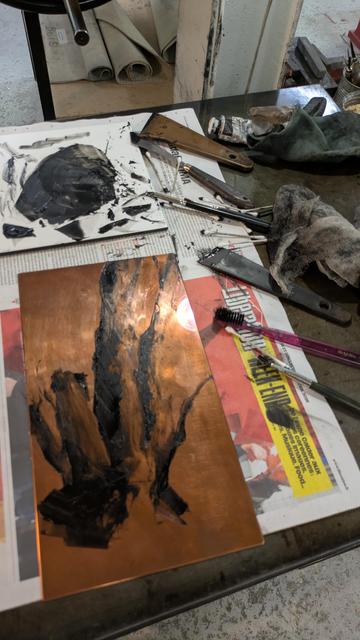 Plaque de cuivre avec dessin à l'encre noire en cours, sur un plan de travail encombrés de spatules, pinceaux, cotons tiges, etc.