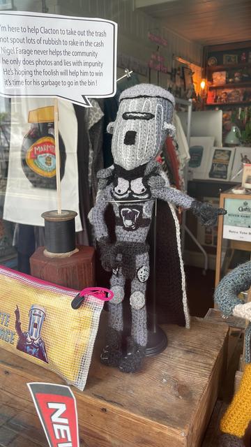 A crocheted and needle-felted figure, about 35cm high, in grey and black with a distinctive large space helmet and cape stands in a shop window. It is British national treasure, political saviour and all round hero COUNT BINFACE! A speech bubble coming out of his mouth reads: “I’m here to help Clacton to take out the trash / not spout lots of rubbish to rake in the cash / Nigel Farage never helps the community / he only does photos and lies with impunity / He’s hoping the foolish will help him to win / it’s time for his garbage to go in the bin!”