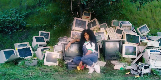 wallpaper which is an extended version of cover of the album "ctrl" by sza. it shows several old computer parts, mostly beige but some black screens and keyboards, piled up on a meadow under a tree. in front of the monitors there's a person (sza) leaning on and resting next to one of the monitors, looking at the camera