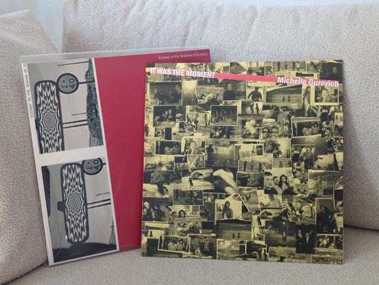 Two vinyl records sitting on a sofa, overlapping each other. In the front, "It was the moment" album. Cover art is lots of small photos of moments from the life of Michelle Gurevich, with a red strip at the top for the album name. Behind, the album "Ecstasy in the shadow of ecstasy". Cover art is on the left two black and white photos of a car's rearview mirror reflecting a hypnotic, wavy optical illusion pattern, in a rural area, stacked photos. The right side is a solid red color.
