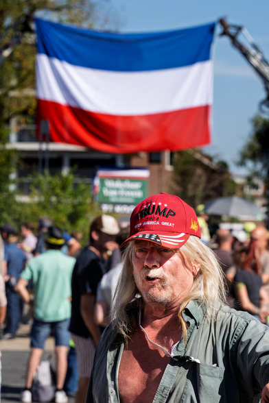 Foto van een man met lang haar en een rood Maga Trump-petje op, lange, blonde haren, baard en snor, open groen overhemd. Op de achtergrond een omgekeerde Nederlandse vlag en nog wat demonstranten.
