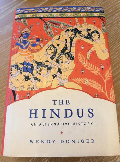 A photograph of the front cover of a book. The top two-thirds features an illustration in a traditional Indian miniature painting style on an orange background, depicting several figures with blue and light skin tones intertwined among green leaves and gold floral motifs, beneath a blue and white patterned border. The bottom third is off-white and contains text in blue and black capital letters. The text reads: "THE HINDUS AN ALTERNATIVE HISTORY", followed by a small red four-pointed star, then "WENDY DONIGER".