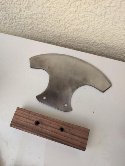 The current state: the polished and sharpened blade (now silver) with two holes where the grip will attach with a elongated square piece of wood with matching holes. The grip has yet to be shaped and attached.