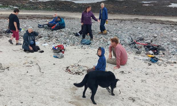 A group of people, some sitting, and some standing, plus a black dog, on a beach. The beach is dry sand in the foreground, pebbly in the middle distance, and seaweed covered rocks in the distance.