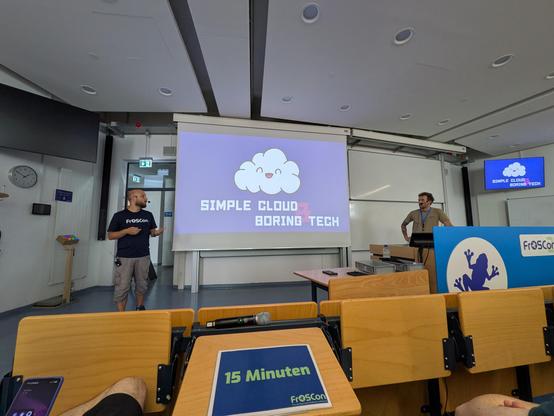 A stage of Froscon room HS7 with Lucas and Dirk on stage, the slide showing a slide of a jolly cloud with the title "simple cloud and boring tech"