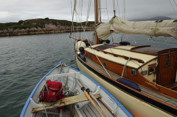 A dinghy with blue gunwales and pale grey interior is alongside a wee cream and wood ketch under a grey sky with a rocky pink granite coast behind.