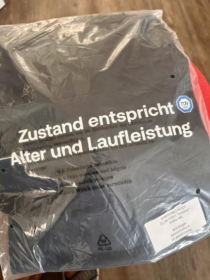 T-Shirt in Verpackung. Der Text auf dem T-Shirt lautet "Zustand entspricht Alter und Laufleistung"