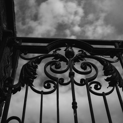 A black and white photograph of a wrought iron gate taken from below looking above. The gate has fancy curlicues and behind there is a cloudy sky.