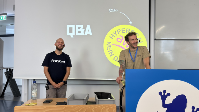 Lucas Dohmen (links) und Dirk Breuer (rechts) vor einer Leinwand auf der Q&A steht. Beide lachen. Vor ihnen steht ein Tisch und ein Rednerpult.