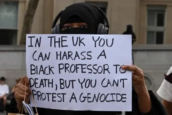 An attendee at last night's memorial for Jason Arday in Trafalgar Square holds a white placard reading IN THE UK YOU CAN HARASS A BLACK PROFESSOR TO DEATH, BUT YOU CAN'T PROTEST A GENOCIDE.