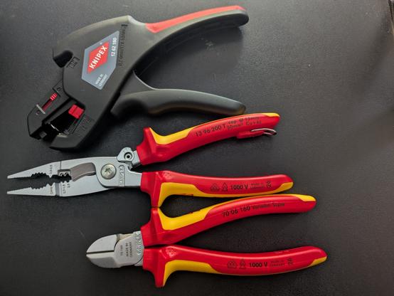 Three hand tools sit on a chair: 
- Knipex auto cable striper 19 62 180
- Knipex electrical installation pliers, which in addition to being pliers are a Ferule crimper, cable strippers for two sizes of wire, and cable shears. A spring holds them slightly open, to aid one handed use; a catch can be used to choose between fully or partially open. They have attractive Red and Yellow over moulded handles, covered in markings saying they're VDE certified tools with 1000V insulation to support working on live cables (or in this case to give additional protection in case of accidentally live cables). Product number 13 96 200
- Some Knipex side cutters, also with the fancy handles; nr. 70 06 160