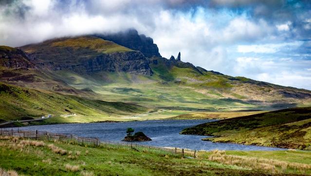A view across a loch to a cloud covered hill with two pinnacles projecting on the skyline. A road runs by the loch with some vehicles on it.