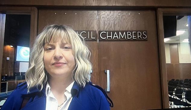Ashley Gjovik wearing a blue jacket and white button down with long-bob length blonde hair looking at the camera in front of the Santa Clara City Council chambers
