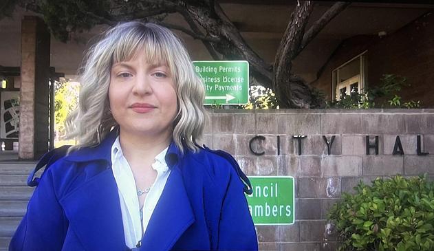 Ashley Gjovik standing confidently in front of the Santa Clara City Hall exterior entrance looking at the camera with long-bob length hair and a turquoise necklace. In the background there is a brick divider with the words "city hall" on it and below it a green and white sign that says "Council Chambers". Its a nice sunny day outside.