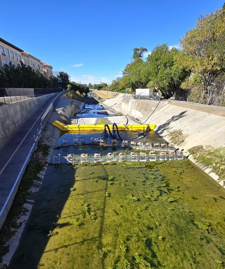 Photo of the San Tomas Aquino/Saratoga Creek from the Scott Blvd overpass in Santa Clara, facing north and showing green murky water, and some sort of construction materials blocking the flow of the creek so it pools before a "drop" downward and two suction machines apparently pulling the water out of the creek and into large black tubes.