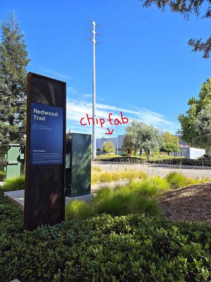 Photo of the chip fab at 3250 Scott Blvd behind a placard from City of Santa Clara for a stop on the Redwood Trail, part of a city park. The photo shows plants and landscaping along a nature trail and no signage or warnings on the chip fab building. Ashley annotated in red and with an arrow where the stealth chip fab is in the photo, as described.