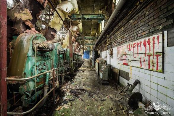 An abandoned industrial interior features large, decaying green machinery lining one side and a tiled wall with red markings on the other, debris covering the floor.