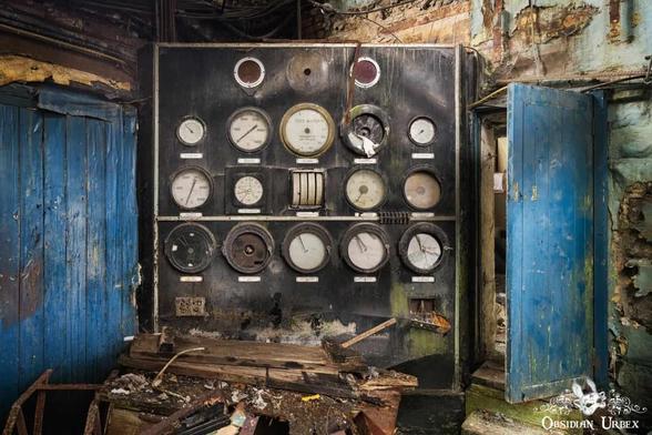 An old, dark control panel filled with many circular gauges and dials. It stands in a derelict room with peeling blue wooden doors