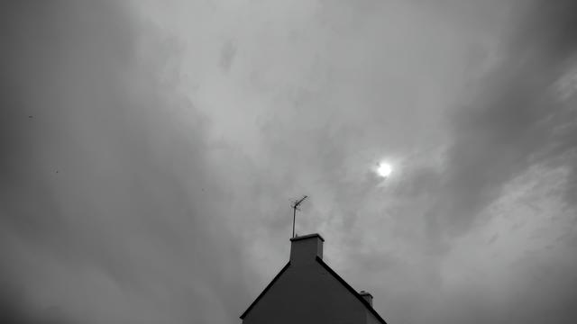 Une image en noir et blanc montrant le toit d'une maison avec une cheminée et une antenne. Le ciel est couvert de nuages, et une petite partie du soleil est visible parmi les nuages.