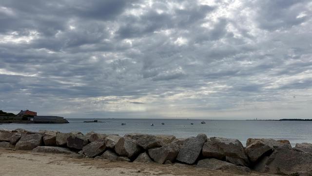 Une scène côtière représentant une plage de pierre avec de gros rochers au premier plan, des eaux calmes au milieu et un ciel rempli de nuages gris. Quelques bateaux sont visibles sur l'eau, et un rivage lointain peut être vu en arrière-plan.
