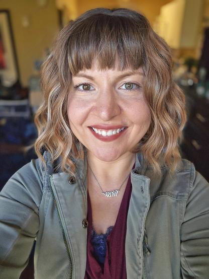 Profile photograph of Ashley with her facing the camera and smiling with her hair in a light brown long-bob hairstyle, a blue lace camisole with a maroon drapey tank, a light army green jacket, and a necklace with crystals in the shape of a branch with leaves.