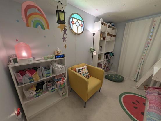 Photo of a children’s bedroom. On the left side of the room is a half-height bookshelf with clothes baskets; on top is a salt lamp, clock, and tissues; above hang a rainbow craft, stars, and paper lantern craft. In the center of the room is a yellow chair with a Christian Robinson pillow with little illustrated children; above on the wall is a large circular light that looks like the stained glass window in Bluey and Bingo’s bedroom; next to the chair is a black floor lamp. On the right side of the room is a large, floor-to-ceiling bookshelf with stuffed animals, clothes, and books; in front of the shelf is a large decorative leaf pillow with illustrated bugs. On the ceiling and walls are glow-in-the-dark stars.