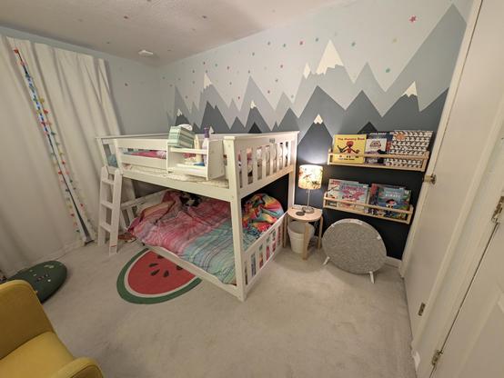 Another angle of the room, facing the opposite wall. There are white wooden bunk beds on the left with rainbow blankets. A half-circle watermelon rug is pushed up against the bunk bed base. To the right is a small table with a lamp on top and trash can underneath. On the wall are two shallow shelves displaying library books, above an IKEA air purifier. The back wall is painted to look like a mountain range, with glow-in-the-dark stars all over the sky portion and the ceiling.